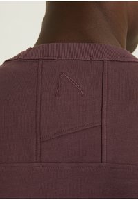 Felpa bordeaux con scollatura a costine, caratterizzata da cuciture uniche e una forma ricamata distintiva sul retro. Tessuto morbido e texture piacevole.