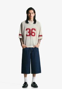 T-shirt en jersey crème à manches courtes avec des rayures rouges et le numéro brodé 36, associé à un short en denim taille large bleu foncé et des chaussures noires.