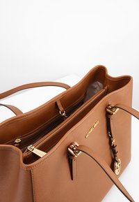 Sac fourre-tout en cuir marron avec ferrures dorées, doté d'une poche zippée et de compartiments intérieurs. Texture lisse et forme structurée.