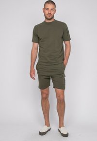 Ensemble en mélange de coton vert olive, comprenant un t-shirt à manches courtes et un short, avec col rond et poche cargo sur le short, associé à des chaussures blanches à enfiler.