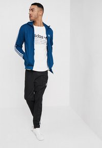 Modrá sportovní bunda se bílými pruhy na rukávech, bílá tričko s černým logem, černé kalhoty s bílými pruhy, bílé tenisky.