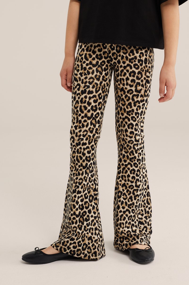 Enfant portant un pantalon évasé à imprimé léopard, des ballerines noires et un t-shirt noir, debout sur un fond neutre.