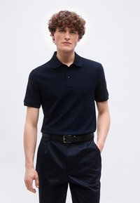 Νεαρός με σγουρά μαλλιά, φορώντας πολο σε σκούρο μπλε ναυτικό χρώμα με υφή και ταιριαστό παντελόνι με μαύρη ζώνη, στέκεται με το ένα χέρι στην τσέπη.