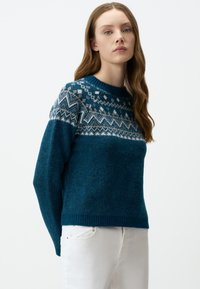 Teal gebreide trui met een gebloemd yoke in lichtblauw en wit, ronde hals, geribde mouwen en zoom. Draagt met witte jeans.