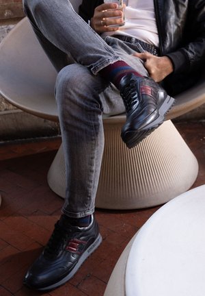 Black leather sneakers with red accents, textured upper, padded collar, rubber sole, worn with gray denim and patterned socks, seated on a sculptural chair.
