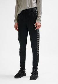 Svarta träningsbyxor med avsmalnande skärning, sidochevronsmönster i mörkgrått och elastisk midja. Tillverkade av ett mjukt, lätt material.