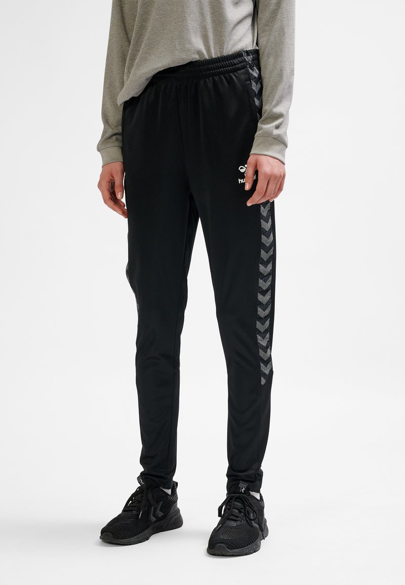 Svarta träningsbyxor med avsmalnande skärning, sidochevronsmönster i mörkgrått och elastisk midja. Tillverkade av ett mjukt, lätt material.