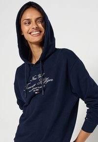 Felpa con cappuccio color blu navy, dotata di tasca a marsupio, cappuccio con coulisse e logo ricamato in crema. Presenta polsini a costine e una texture morbida.