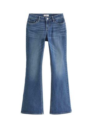 Teenagepige, der står med den ene hånd på hoften iført hvid t-shirt, blå jeans og hvide sneakers mod en ensfarvet baggrund.