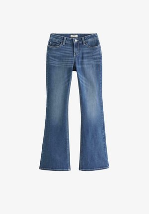 Teenagepige, der står med den ene hånd på hoften iført hvid t-shirt, blå jeans og hvide sneakers mod en ensfarvet baggrund.