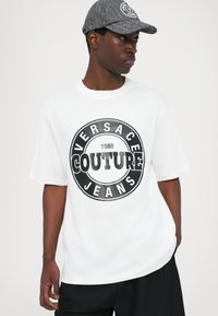 Balta didelė marškinė su dideliu juodu apvaliu logotipu, pažymėtu "VERSACE COUTURE JEANS 1988." Lygus audinys, apvalus iškirpimas, trumpomis rankovėmis.