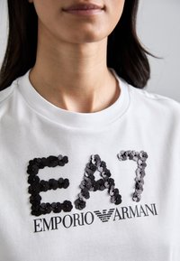 Bijela pamučna majica s izvezenim crnim šljokicama "E A 7" i ispisanim crnim tekstom "EMPORIO ARMANI", dizajn s okruglim izrezom i glatkom teksturom.