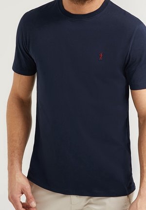 Homme portant un t-shirt bleu marine à manches courtes avec un petit logo rouge brodé sur le côté gauche de la poitrine.