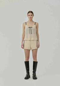 Conjunto de dois peças em renda na cor creme, apresentando um topo peplum com laço preto e calções com detalhes laterais. A modelo usa botas altas pretas.