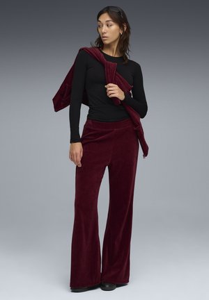 Burgundy corduroy-materiaalin leven pantsit, joissa on samanvärinen huivi, yhdistettynä istuvaan mustaan pitkähihapaitaan. Sileä pinta, rento istuvuus ja hillitty kiilto.