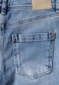 Svetle modré denimové džíny s zadní kapsou se jednoduchým švem a kovovým knoflíkem jako ozdobou. Štítek přišity na pásku.