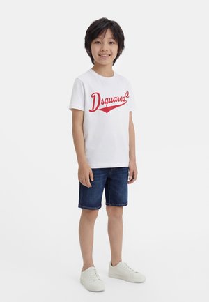 Ένα χαμογελαστό αγόρι στέκεται φορώντας λευκό T-shirt Dsquared2 με κόκκινο λογότυπο, σκούρα τζιν σορτς και λευκά αθλητικά παπούτσια, σε απλό λευκό φόντο.