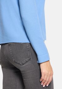 Jasnoniebieski sweter z prążkowanym kołnierzem i mankietami, w połączeniu z ciemnoszarymi dopasowanymi dżinsami. Gładka tekstura i swobodny design.