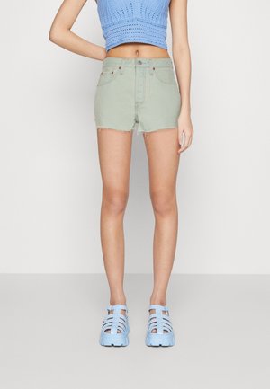 Femme portant un short en denim vert clair, un crop top en maille bleu et des sandales compensées bleu clair, debout devant un fond uni.
