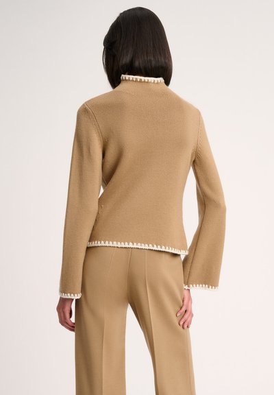 Pull en maille beige avec des manches longues et un col montant, orné de bordures festonnées blanches et d'une coupe décontractée.