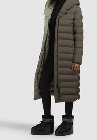 Langer, gesteppter Mantel in Olivgrün und Khaki, mit Kapuze, Druckknöpfen und seitlichen Reißverschlusstaschen, kombiniert mit schwarzen, knöchelhohen Stiefeln.