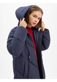 Doudoune bleu marine avec capuche, présentant un design matelassé, une fermeture éclair et des poches latérales. Portée par-dessus un pull rouge à col montant.