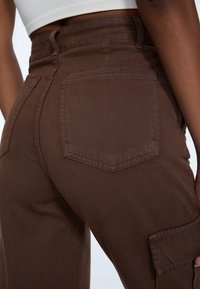 Bruine cargo broek met een hoge taille, gemaakt van een textuurstof, met patchzakken en een ontspannen pasvorm. Zichtbare stiksels en details op de zakken.