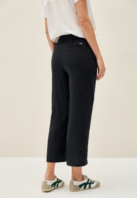 Modella femminile che mostra la vista posteriore, indossando una maglietta bianca e pantaloni neri texturizzati abbinati a sneaker casual su uno sfondo neutro.