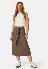 Gonna marrone di media lunghezza con bottoni frontali, dettaglio della vita con lacci e due grandi tasche. Indossata con un top cropped bianco e sneaker bianche.