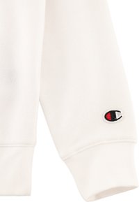 Material de sudadera blanca con puños acanalados y un pequeño logo bordado con acentos rojos y negros en la manga. Textura suave.
