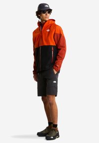 Chaqueta en bloques de color naranja, negro y rojo; con cremalleras, combinada con pantalones cortos negros a juego y zapatos de senderismo oscuros.