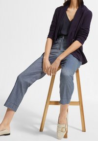 Granatowa marynarka, ciemna bluzka i jasnoniebieskie spodnie w fasonie cropped z delikatną teksturą. Beżowe buty typu slip-on z okrąłym złotym akcentem. Drewniany taboret.