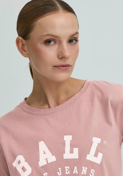 Mladá žena s odkrytými vlasmi, oblečená v prašnej ružovej tričku s nápisom "BALL JEANS" v veľkých bielych písmenách naprieč hrudníkom.