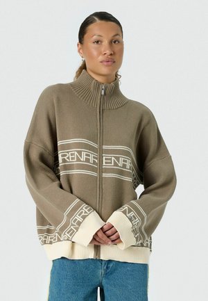 Nő bézs színű, cipzáras pulóvert visel fehér szövegmintákkal és kék farmerral, lábait összekulcsolva áll sima háttér előtt.