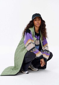 Grüner, gepolsterter langer Weste über einem mehrfarbigen Strickpullover mit lila und schwarzen Streifen, kombiniert mit dunklen Jeans und silbernen Sneakers.