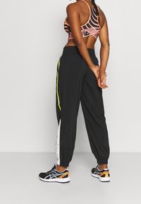 Žena s copánky, oděná v černých sportovních kalhotách s žlutým a bílým pruhem a černých běžeckých botách, stojící s rukama za zády.