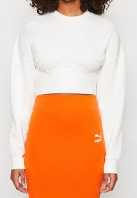 Felpa bianca tagliata con orlo arrotondato e maniche lunghe, abbinata a una gonna aderente di un vivace arancione con logo Puma bianco.