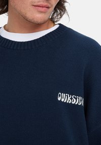 Suéter de malha azul marinho com decote canelado e logótipo bordado em branco 'QUIKSILVER' na parte da frente, com um acabamento texturizado.