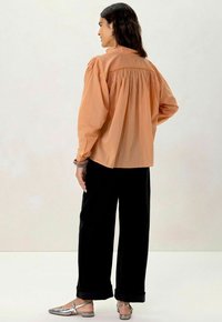 Blouse oversize de couleur rouille avec détail froncé au dos, manches longues, et pantalon noir. Le modèle porte des chaussures plates métalliques.