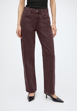 Femme portant un jean violet foncé taille haute, jambes droites, un haut noir et des talons noirs pointus sur fond blanc.