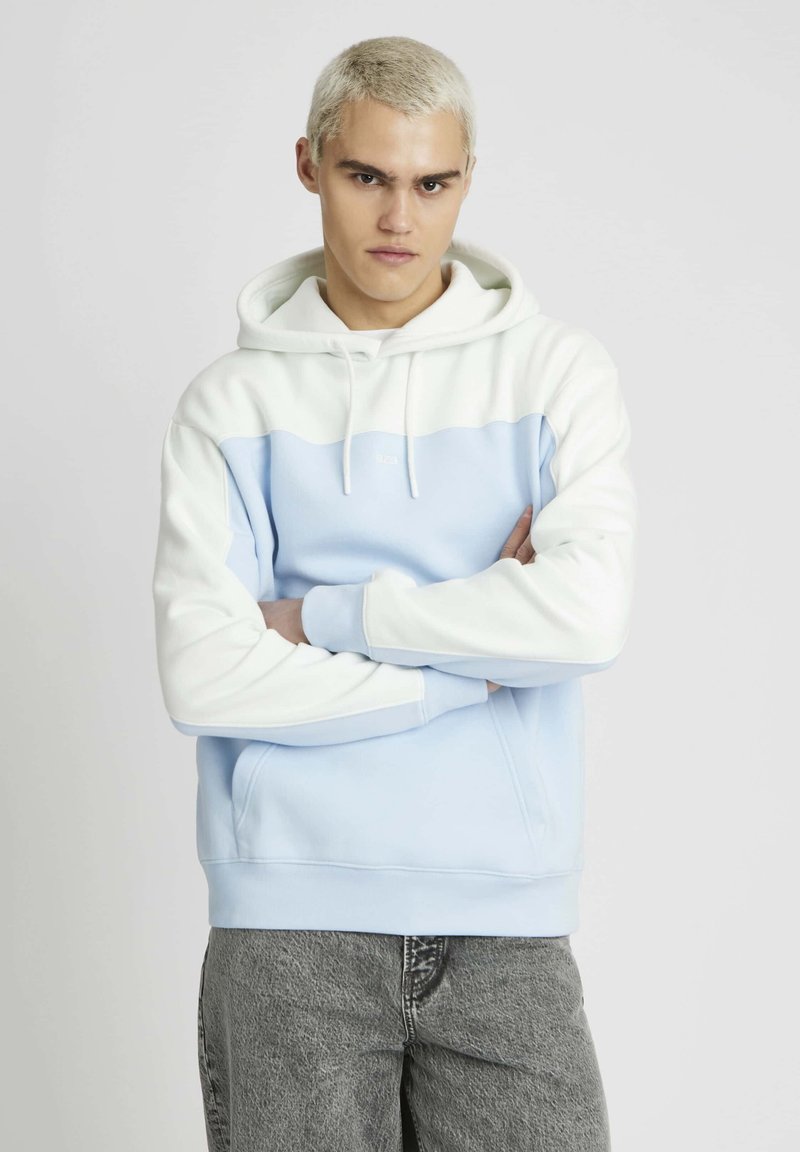 Haut à capuche bleu clair avec des accents blancs. Comprend une poche kangourou, une capuche avec cordon de serrage et des poignets côtelés. Texture lisse et design décontracté.