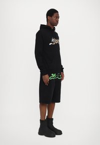 Sweat à capuche noir avec un design imprimé léopard et une poche frontale, associé à un short noir avec des accents vert citron, et des bottes noires à chevilles.