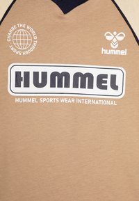 Beżowa odzież sportowa z czarnym wykończeniem, z białym logo "hummel", emblematem pszczoły oraz grafiką globu z napisem "Zmieniać świat przez sport".