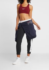 Reggiseno sportivo Nike bordeaux, felpa FILA blu navy legata in vita, pantaloni neri tapered con strisce bianche, sneakers bianche.
