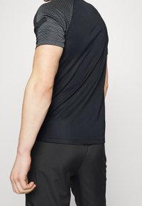 Camisa atlética de manga corta en negro con detalles texturizados en gris en los hombros. Tejido ligero, diseño ajustado, textura suave, estilo minimalista.