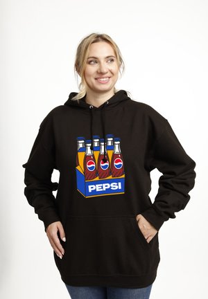Smilende kvinde iført en sort hættetrøje med et farverigt grafisk motiv af seks Pepsi-flasker i en blå carrier på forsiden, stående med hænderne i lommen.