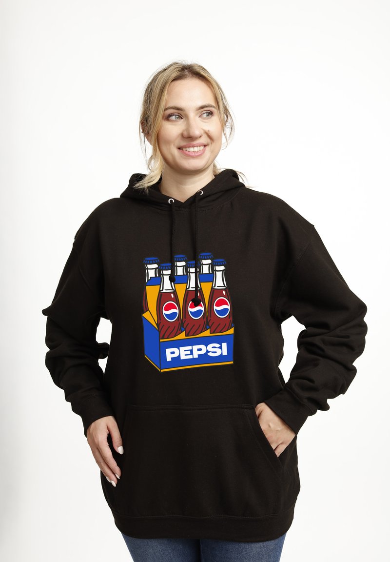 Donna sorridente che indossa un felpa nera con una grafica colorata di sei bottiglie di Pepsi in un portabottiglie blu sul davanti, in piedi con le mani nelle tasche.