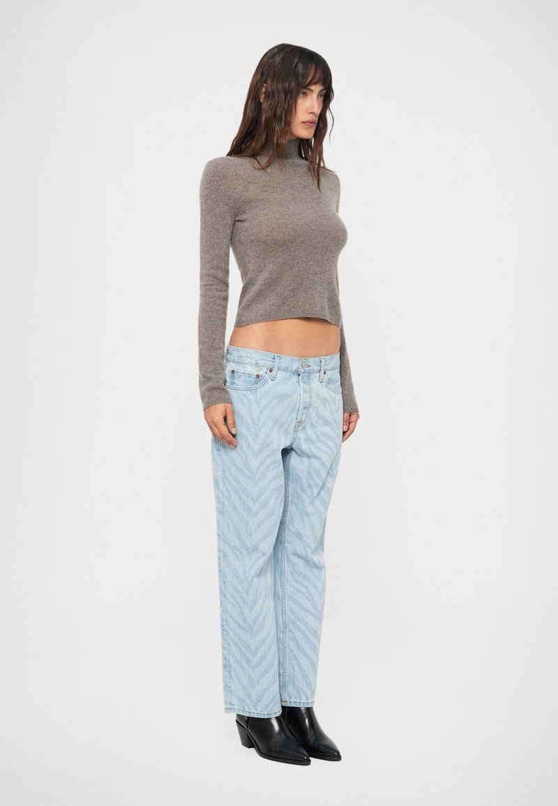 Vrouw staat in een grijze langemouwen cropped sweater, lichtblauwe gedessineerde jeans en zwarte puntige enkelboots tegen een witte achtergrond.