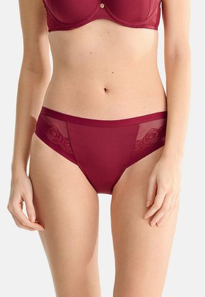 Vrouw die een bijpassende donkerrode beha en slip draagt met doorschijnende kanten details aan de zijkanten van de slip.