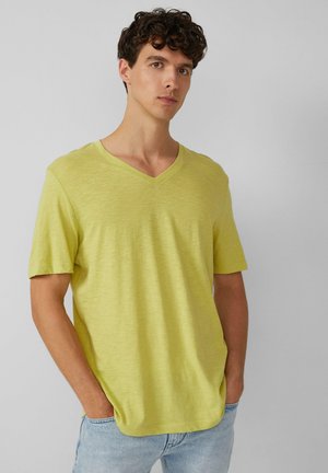 Jasnożółta koszulka typu V-neck z krótkim rękawem, wykonana z miękkiego materiału. Gładka tekstura i luźny krój, noszona z jasnoniebieskimi dżinsami.